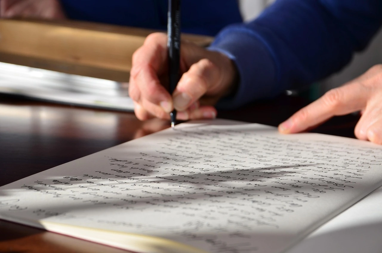 Close-up of a person writing in cursive on paper with a pen, while another hand points at the page.