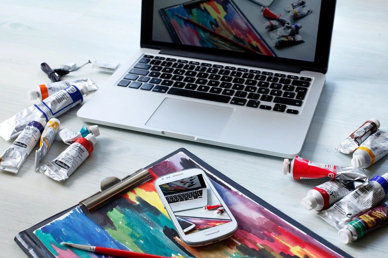 Colorful art supplies, a laptop, and a smartphone displaying digital art on a wooden desk.